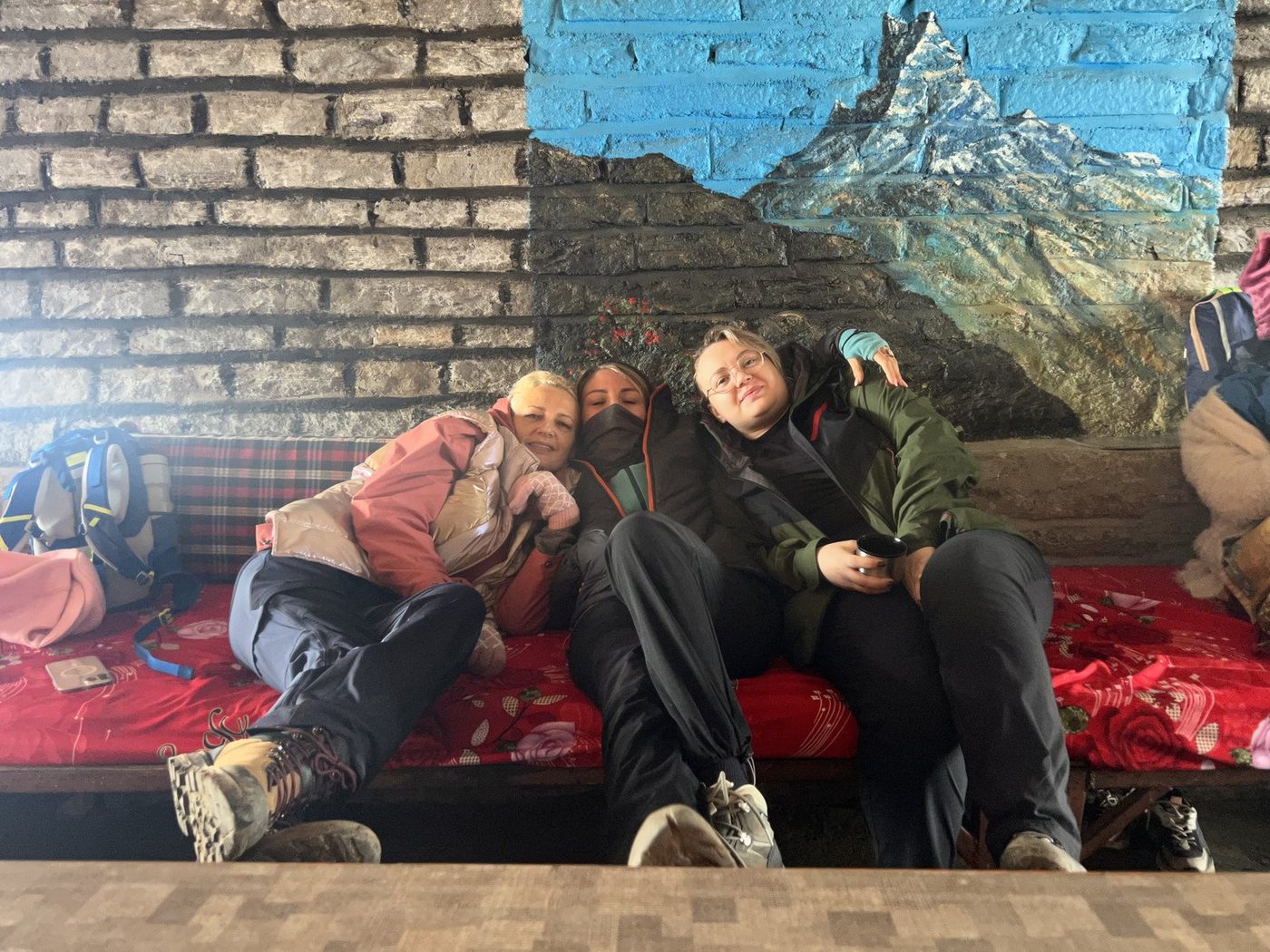 Three tired trekkers resting on a red bench below a painted mural of a snow peak