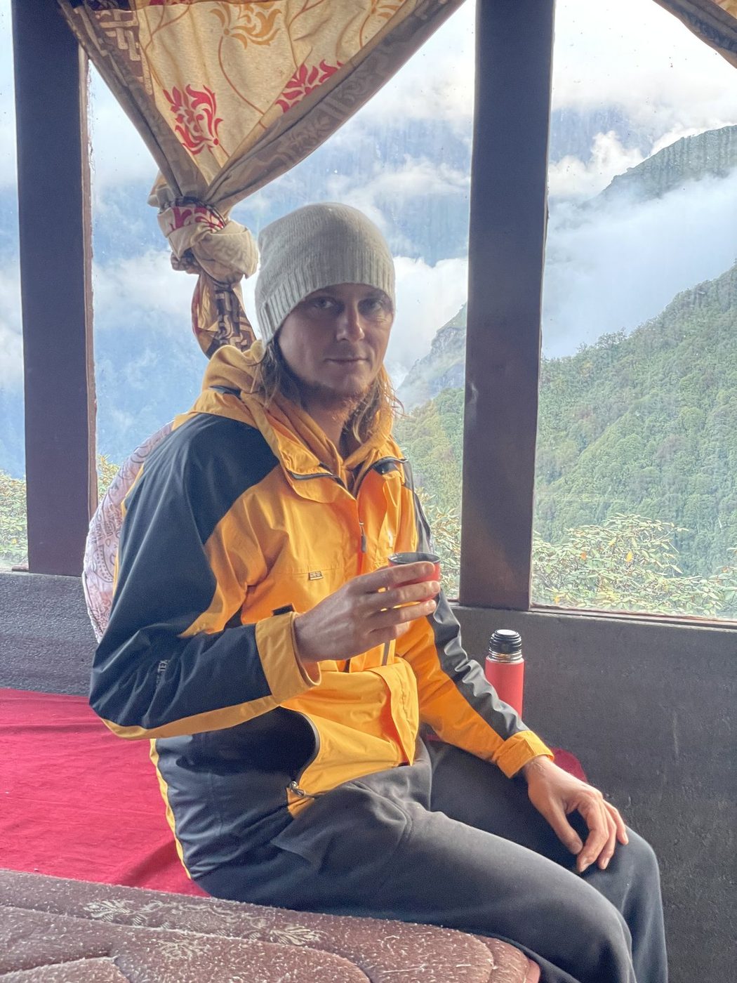 A trekker in a yellow jacket and wool hat holding a small cup of tea, with misty mountains behind