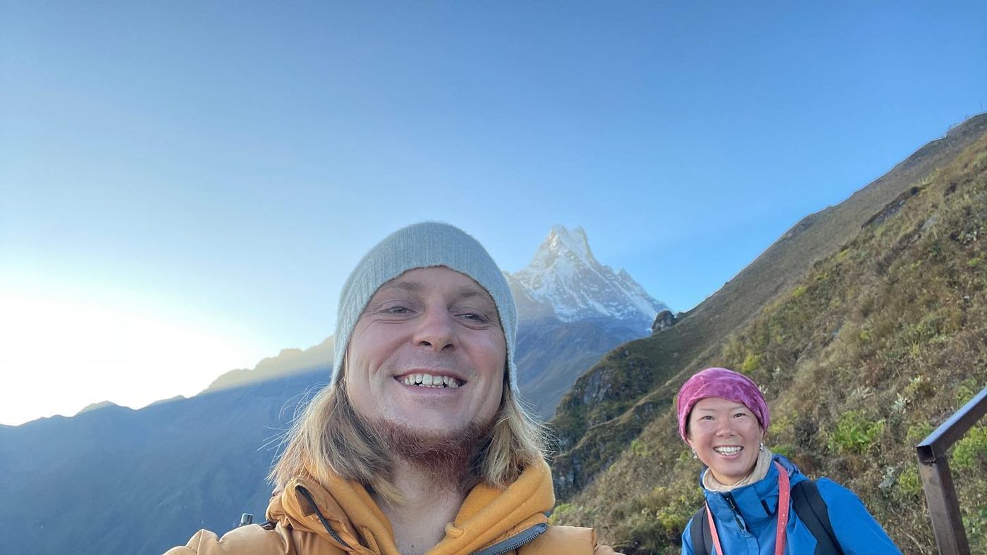 Two smiling trekkers in a selfie with a snow peak and the morning sun behind them