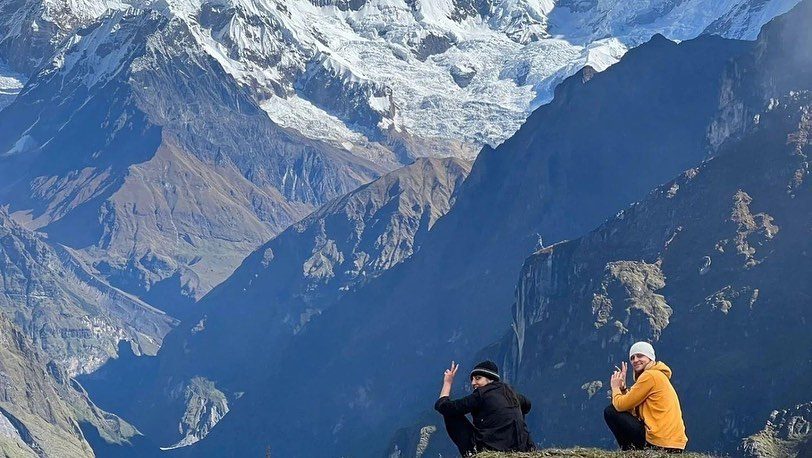 Two trekkers sitting on a ridge and waving, with a huge wall of snow mountains behind them