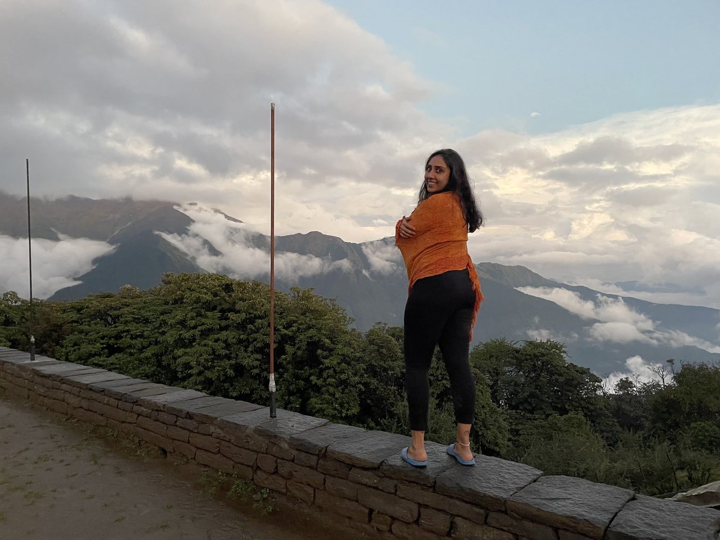 A woman in an orange top standing on a stone wall looking back, with hills and clouds in evening light