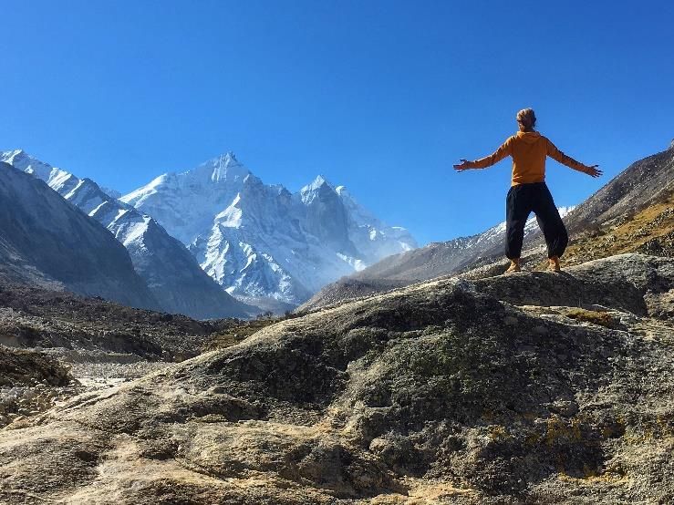 A person standing on rocks with arms open, facing snow mountains