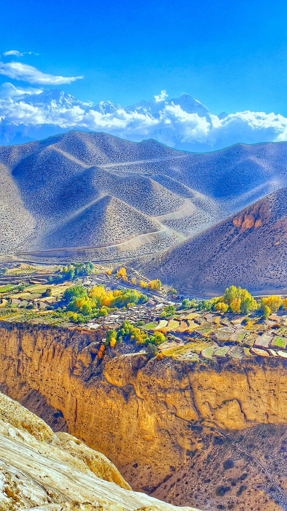 A village with yellow trees at the edge of a cliff, with brown hills and snow peaks beyond
